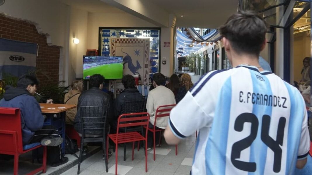 Interior de un bar en Rosario con pantalla gigante mostrando un partido de fútbol, con pocos clientes