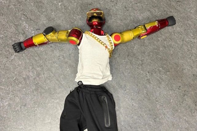 Robot rojo y dorado tirado en el suelo con camiseta blanca, pantalones negros, cadena dorada, anteojos de sol y bigote falso